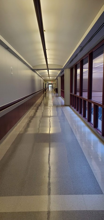 A seemingly endless hallway in a hospital. The floors have a shine to them and the yellow tint to the lighting makes it feel older than it probably is.