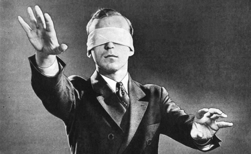 Old black and white photo of blindfolded man in a suit reaching out in front of him.